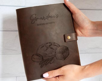 May include: A brown leather-bound recipe book with the words "Grandma's BAKING SECRETS" engraved on the cover. The cover also features an illustration of bread and wheat. The book is held by a person.
