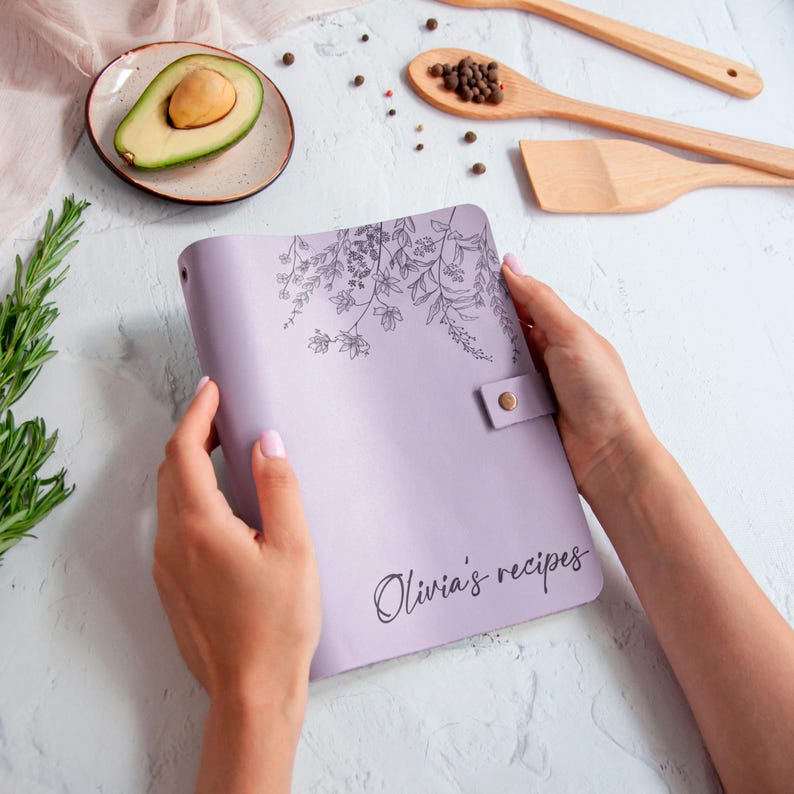 Lavender personalized leather recipe book with floral engraving, held in hands on a styled kitchen flat lay with avocado and wooden utensils.