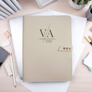 Op de afbeelding: Een beige rechthoekige notitieboekhoes met een drukknoopsluiting voorzien van drie metalen studs. De voorzijde is bedrukt met de letters VA, een diagonale lijn en de naam VIOLET ALEXIS in een donkergrijs lettertype, gecentreerd op het oppervlak.