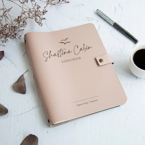 Può includere: Un libro degli ospiti rosa pallido con la scritta "Shastina Cabin Guest Book" e una grafica di uccelli. Il libro ha una copertina in pelle e una chiusura a scatto. Una penna e una tazza di caffè sono accanto al libro.