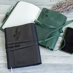 May include: Two leather-bound journals with floral designs engraved on the covers. The journal on the left is black with the name "Marnie" engraved on the front. The journal on the right is green with a floral design.
