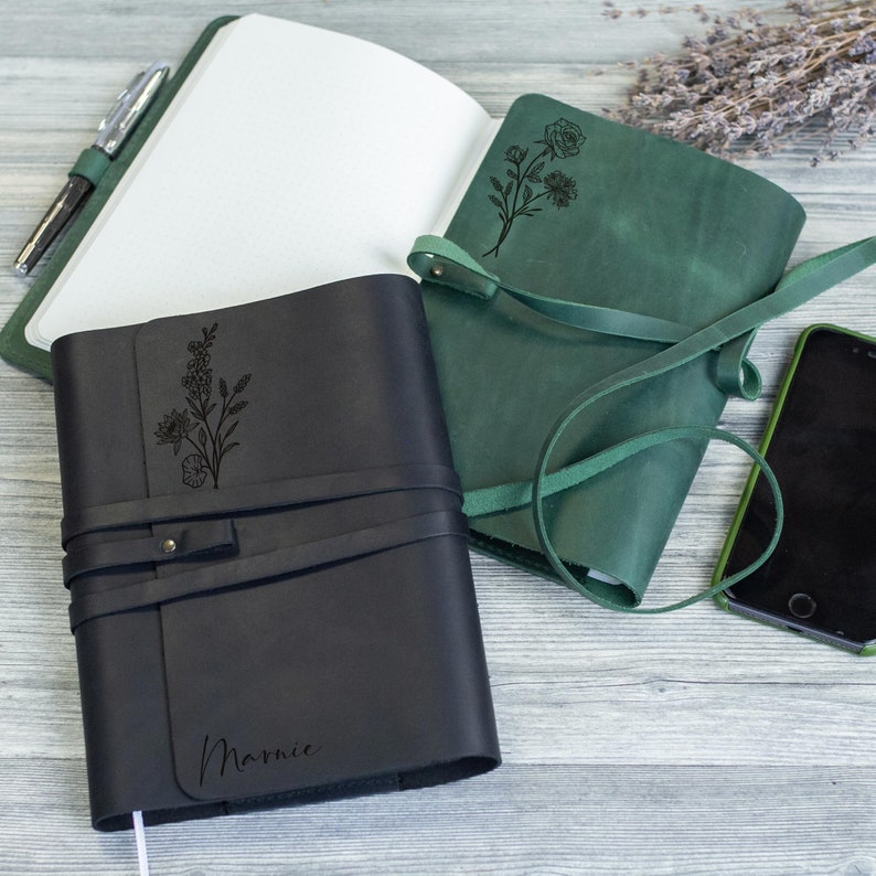 May include: Two leather-bound journals with floral designs engraved on the covers. The journal on the left is black with the name "Marnie" engraved on the front. The journal on the right is green with a floral design.