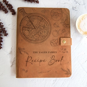 Könnte beinhalten: Ein braunes Leder-Rezeptbuch mit einer Pizza-Illustration und dem Text "THE YAGER FAMILY Recipe Book". Das Cover zeigt eine Pizza, Gemüse und eine Käse-Illustration. Ein Lederriemen mit einem goldenen Knopf sichert das Buch.