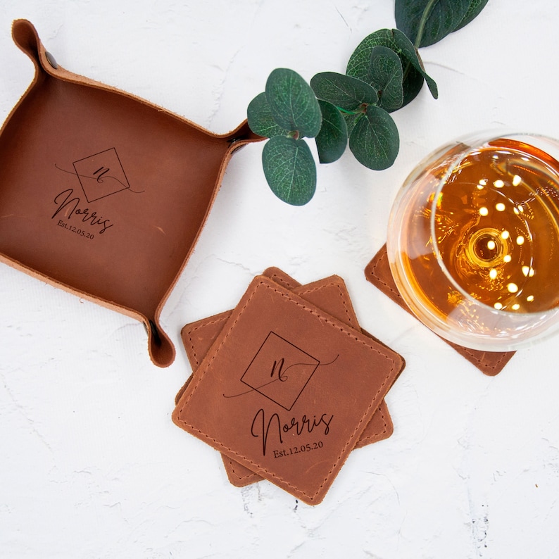 May include: Set of four brown leather coasters with a square design and the text "Norris Est. 12.05.20" in a script font. The coasters are stacked on top of each other. A brown leather tray with the same design and text is also shown in the image.
