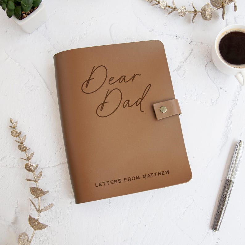 May include: A tan leather-like journal with a snap closure featuring the text Dear Dad in a cursive script and the text LETTERS FROM MATTHEW in a block font. The journal is positioned on a white textured surface with a pen and a cup of coffee.
