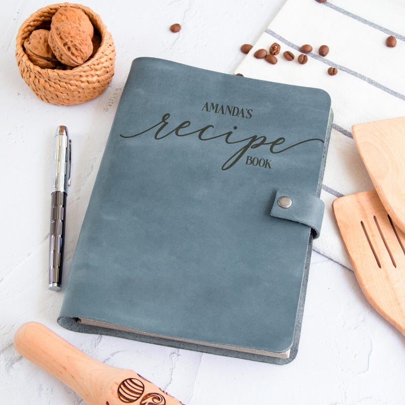 Personalized light blue leather recipe book with engraved custom name, styled with kitchen utensils, walnuts, coffee beans, and rolling pin.
