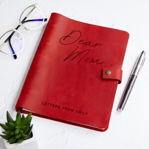 May include: A red leather journal with a snap closure, featuring the text "Dear Mom" in script and "LETTERS FROM EMILY" in block letters. The journal is accompanied by a pair of silver-rimmed eyeglasses and a silver metal pen.