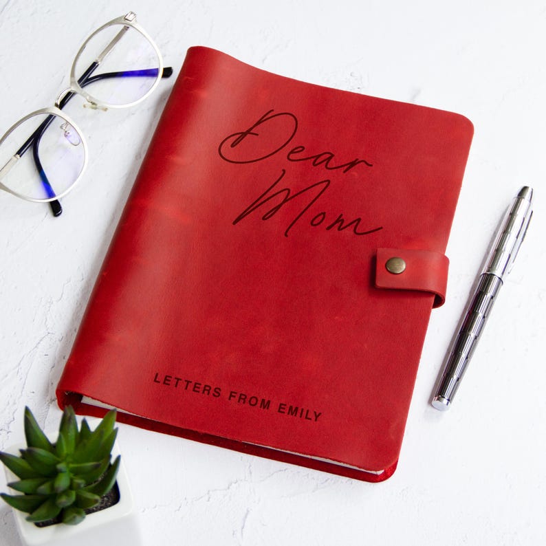 May include: A red leather journal with a snap closure, featuring the text "Dear Mom" in script and "LETTERS FROM EMILY" in block letters. The journal is accompanied by a pair of silver-rimmed eyeglasses and a silver metal pen.