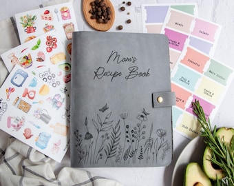 Könnte beinhalten: Ein graues Rezeptbuch mit dem Titel "Mom's Recipe Book" und einem floralen Design. Aufkleber mit Kochillustrationen und Etiketten für verschiedene Lebensmittelkategorien sind ebenfalls vorhanden. Ein Holzlöffel mit Pfefferkörnern und Avocadoscheiben sind ebenfalls sichtbar.