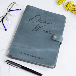 May include: A slate blue leather journal with a snap closure, featuring the text Dear Mom in script and LETTERS FROM EMILY in block letters. The journal is accompanied by a silver metal pen and a pair of silver-rimmed eyeglasses.