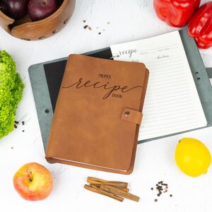 Personalized caramel brown leather recipe book with engraved 'Mom’s Recipe Book' text, open binder pages, and kitchen ingredients on a white background.