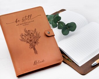 Può includere: Un diario in pelle marrone con le parole "be still and know that I am God" e "Melinda" incise sulla copertina. È presente anche un disegno di bouquet floreale. Il diario è aperto, rivelando pagine a righe.
