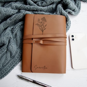 May include: A brown leather journal with a floral design and the name Samantha engraved on the front. The journal has a strap closure and is lying on a white surface.
