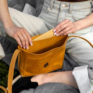 K&ouml;nnte beinhalten: Eine senfgelbe Umh&auml;ngetasche aus Leder mit einem d&uuml;nnen Schulterriemen und einem Frontklappenverschluss mit einem dunklen Metallbeschlag. Der Name Chelsey ist in Schreibschrift auf der Vordertasche der oben offenen Tasche eingepr&auml;gt.