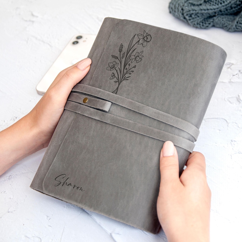 May include: Gray leather journal with a floral design and the name "Sharon" engraved on the front. The journal has a wrap-around closure with a button.