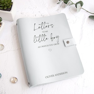 K&ouml;nnte beinhalten: Ein hellgraues Lederjournal mit dem Text "Letters to my little boy as I watch you grow" und dem Namen "Oliver Anderson". Das Journal hat einen Druckknopfverschluss und ist ein liebevolles Geschenk.
