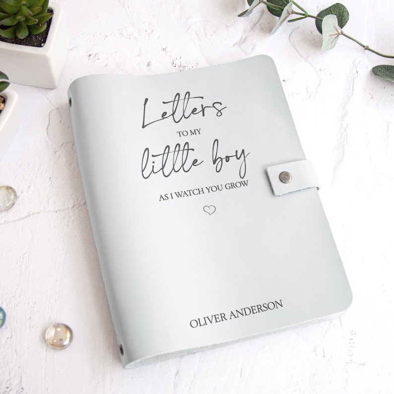 K&ouml;nnte beinhalten: Ein hellgraues Lederjournal mit dem Text "Letters to my little boy as I watch you grow" und dem Namen "Oliver Anderson". Das Journal hat einen Druckknopfverschluss und ist ein liebevolles Geschenk.