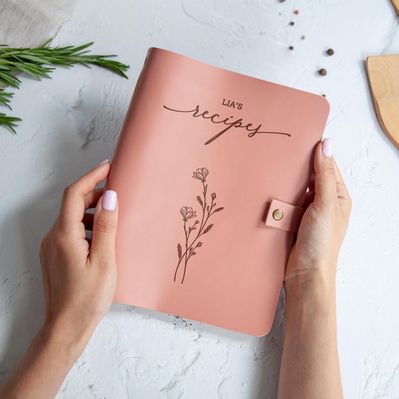 Blush pink personalized recipe book with floral engraving and custom Lia’s recipes text, held in hands on a minimal kitchen background.