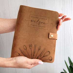 May include: A brown leather-bound recipe book with the text "MELISSA & DAVID WILSON FAMILY RECIPES." The cover features a decorative illustration of kitchen utensils. A leather strap with a snap closure secures the book.
