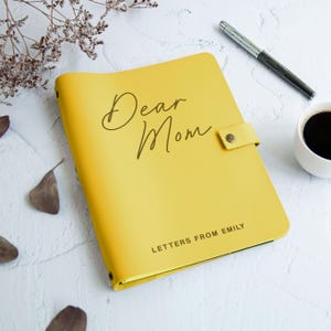 May include: A bright yellow journal with a snap closure, featuring the text Dear Mom in a cursive script and the text LETTERS FROM EMILY in a sans-serif font. The journal is positioned next to a silver pen and a white cup of black coffee.