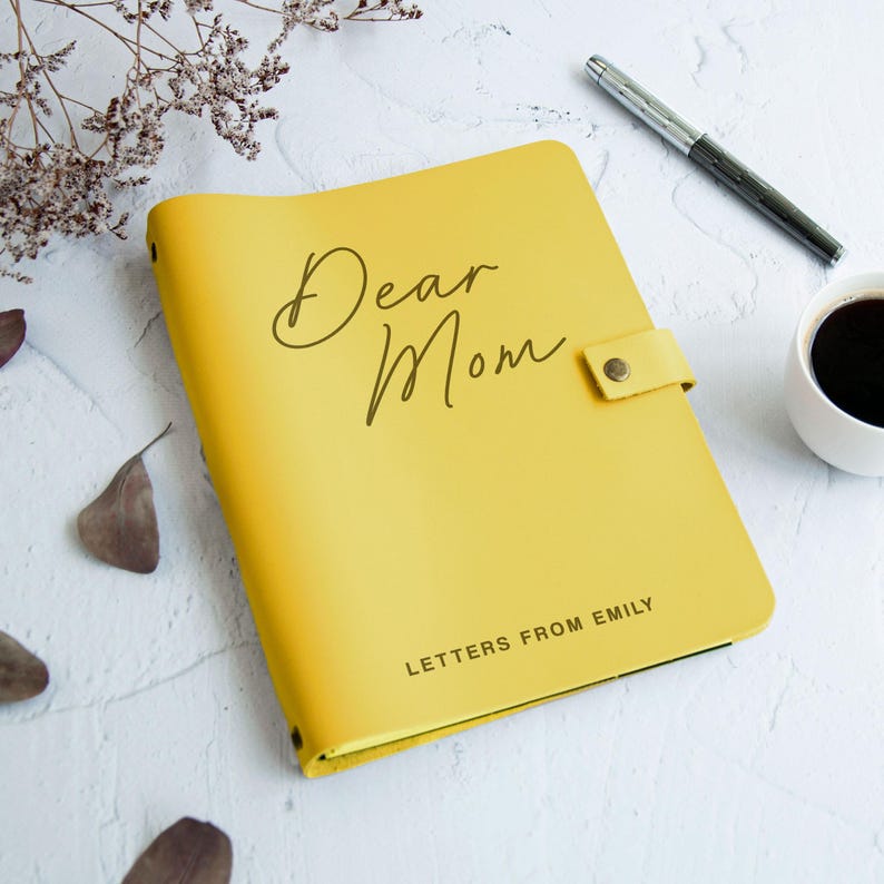May include: A bright yellow journal with a snap closure, featuring the text Dear Mom in a cursive script and the text LETTERS FROM EMILY in a sans-serif font. The journal is positioned next to a silver pen and a white cup of black coffee.
