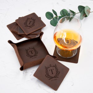 May include: A set of brown leather coasters with a monogrammed shield design and the date "2.17.2022". The coasters are stacked on top of each other and there is a matching leather tray with the same design.