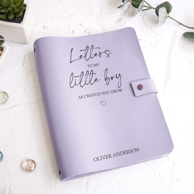 K&ouml;nnte beinhalten: Ein lavendelfarbenes Notizbuch mit dem Text "Letters to my little boy as I watch you grow" und dem Namen "Oliver Anderson" auf dem Einband. Das Notizbuch hat einen Druckknopfverschluss.
