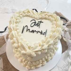 May include: A white heart-shaped cake decorated with white frosting and pearl sprinkles. The cake is inscribed with the words "Just Married" in black icing.
