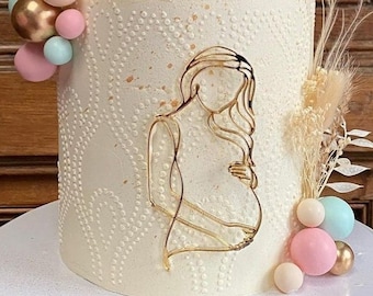 Puede incluir: Un pastel blanco decorado con un contorno dorado de una mujer embarazada. El pastel está cubierto con un grupo de globos de colores pastel y hierba de pampa seca.