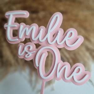 Puede incluir: Topper de pastel rosa y blanco con el texto "Emilie is One" en una fuente cursiva.