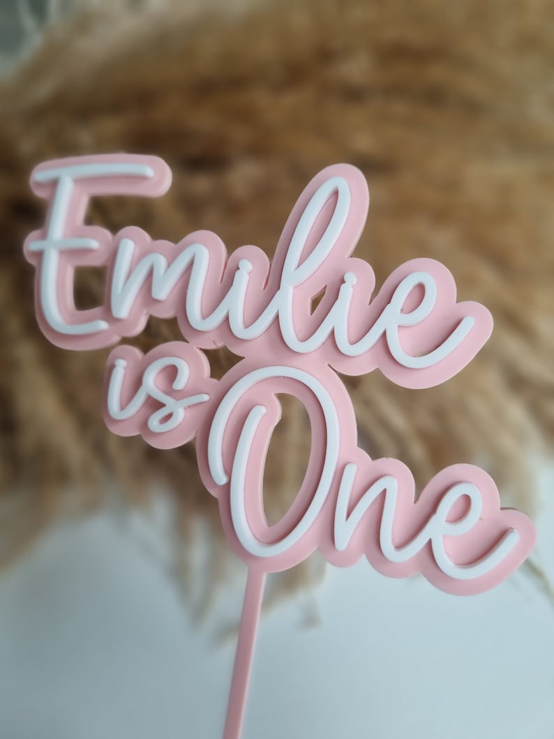 Puede incluir: Topper de pastel rosa y blanco con el texto "Emilie is One" en una fuente cursiva.