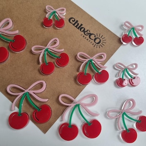 May include: Acrylic cherry charms featuring red cherries, green stems, and pink bows. The charms are arranged on a brown and white surface. The text "Chlo&Co Creates" is visible on the brown surface.