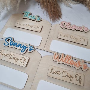 May include: Four wooden signs with blank spaces for writing in. Each sign has a different name written on it in colorful letters: Theo's, Olivia's, Sonny's, and Willow's. The signs also have the words "First Day Of" or "Last Day Of" written on them.