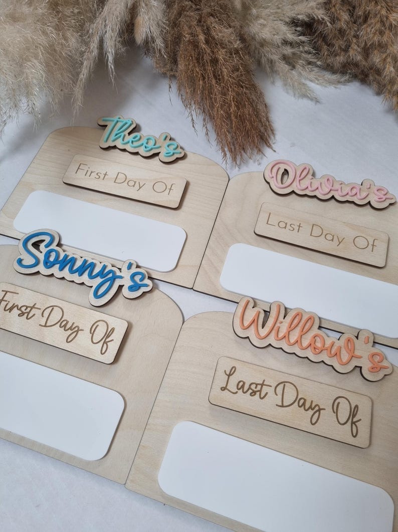 May include: Four wooden signs with blank spaces for writing in. Each sign has a different name written on it in colorful letters: Theo's, Olivia's, Sonny's, and Willow's. The signs also have the words "First Day Of" or "Last Day Of" written on them.