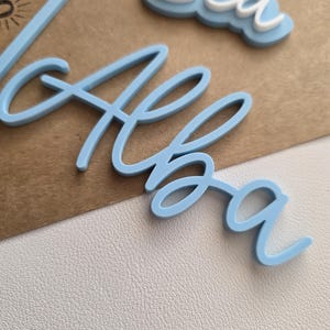 May include: A light blue acrylic cake topper featuring the name Alba in a cursive script font, positioned above a partial word that appears to be Sea in white and light blue layered lettering, with a thin stem extending from the name.
