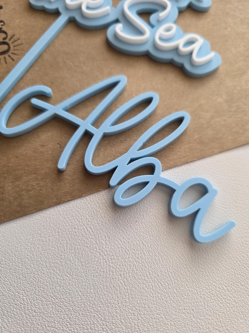 May include: A light blue acrylic cake topper featuring the name Alba in a cursive script font, positioned above a partial word that appears to be Sea in white and light blue layered lettering, with a thin stem extending from the name.