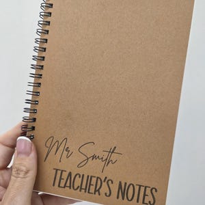 May include: A spiral-bound notebook with a kraft paper cover. The cover features the text "Mr Smith TEACHER'S NOTES" in black script and block letters. The notebook is held in a person's hand.