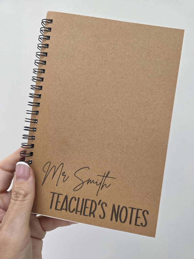 May include: A spiral-bound notebook with a kraft paper cover. The cover features the text "Mr Smith TEACHER'S NOTES" in black script and block letters. The notebook is held in a person's hand.