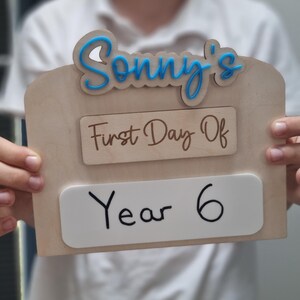 May include: A wooden sign with the text "Sonny's First Day Of Year 6" in blue and brown lettering. The sign is held by a person.
