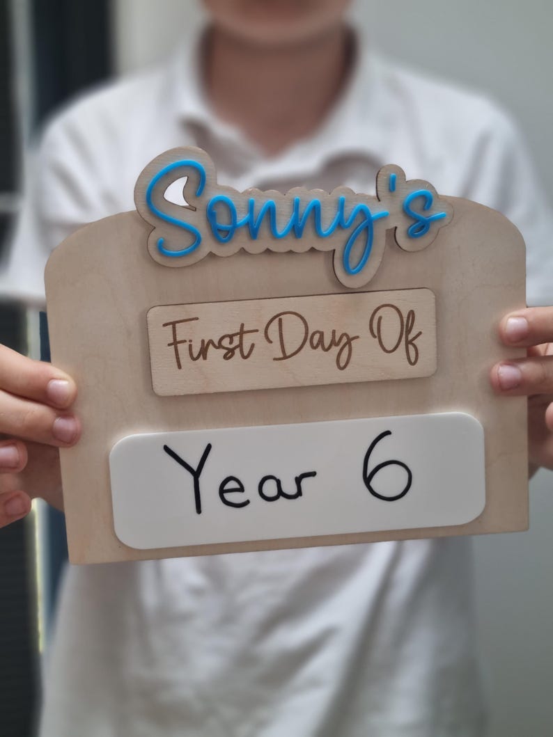 May include: A wooden sign with the text "Sonny's First Day Of Year 6" in blue and brown lettering. The sign is held by a person.