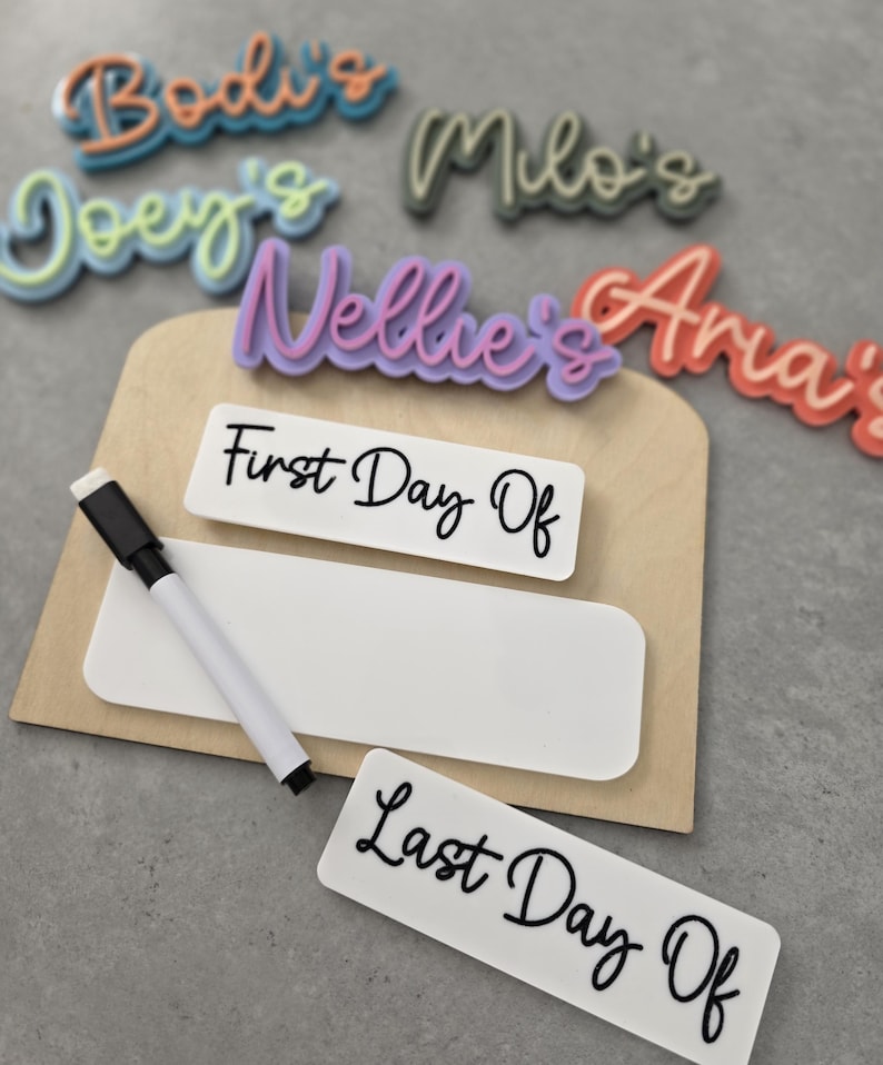 May include: A wooden sign with a dry erase surface and a white marker. The sign has the words "First Day Of" and "Last Day Of" written on it. There are also several colourful name tags with the names "Bodi's", "Joey's", "Milo's", "Nellie's", and "Ana's" written on them.