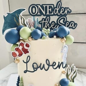 May include: A cylindrical cake with smooth white frosting, decorated with blue and light green spheres, gold balls, and sea-themed cutouts including a stingray, clownfish, seahorse, and turtle. The text reads "ONEder the Sea" and "Lowen".