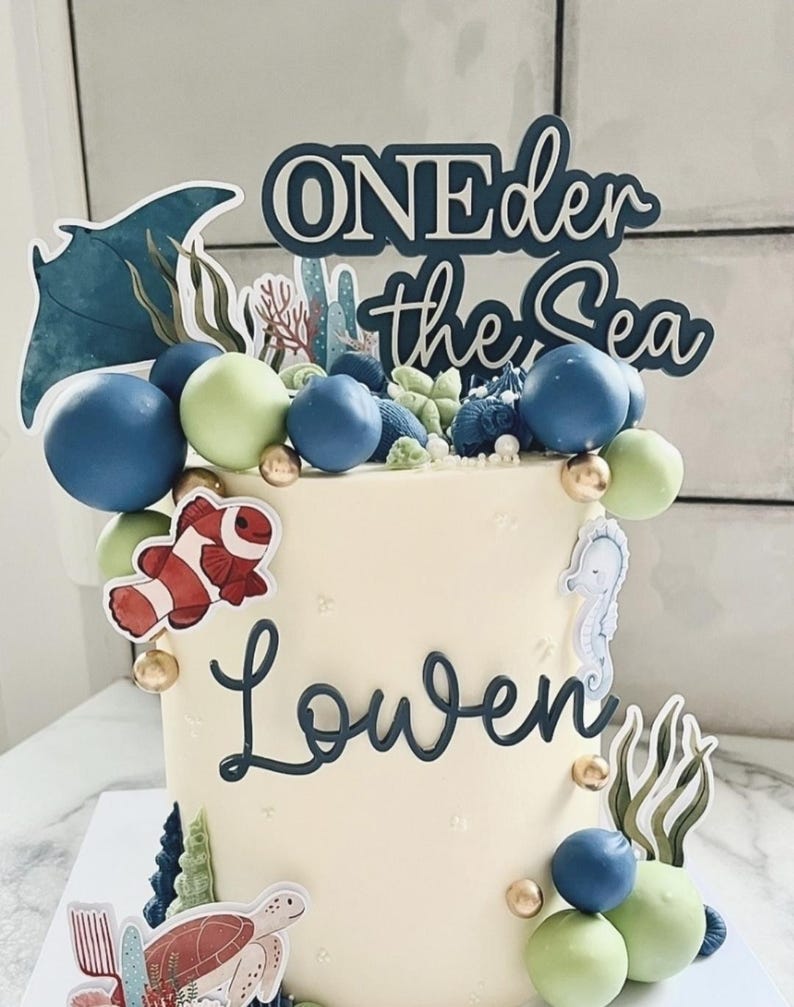May include: A cylindrical cake with smooth white frosting, decorated with blue and light green spheres, gold balls, and sea-themed cutouts including a stingray, clownfish, seahorse, and turtle. The text reads "ONEder the Sea" and "Lowen".