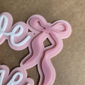 May include: A pink bow-shaped embellishment with the word "love" in white script. The bow has a layered design, and the word "love" is in a cursive font. The background is a light brown color.