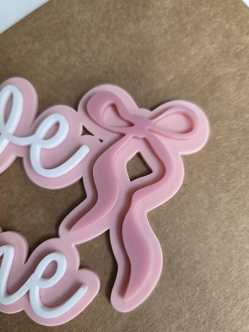 May include: A pink bow-shaped embellishment with the word "love" in white script. The bow has a layered design, and the word "love" is in a cursive font. The background is a light brown color.