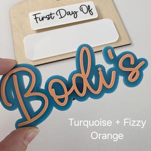 May include: Turquoise and orange acrylic name sign with the text "Bodi's" and the words "First Day Of" on a wooden background.