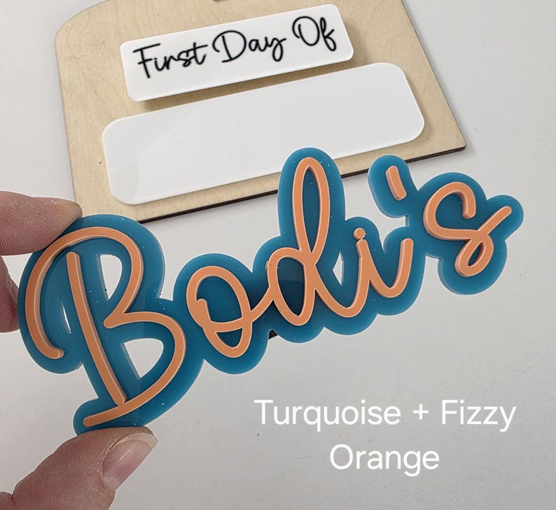 May include: Turquoise and orange acrylic name sign with the text "Bodi's" and the words "First Day Of" on a wooden background.