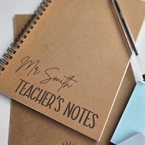 May include: A spiral-bound notebook with a kraft paper cover. The cover is printed with "Mr Smith TEACHER'S NOTES" in black script and block letters. A pen and sticky notes are also visible.