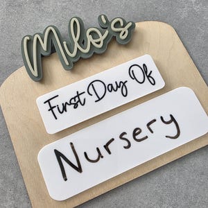 May include: A wooden sign with the name "Milo's" in green and beige letters. The sign also has two white labels, one that says "First Day Of" and the other that says "Nursery".