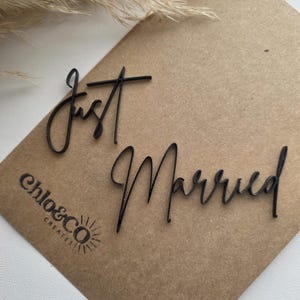 Peut inclure: Mots noirs découpés au laser "Just Married" sur une carte marron. Les mots sont en écriture cursive. La carte porte le logo "chlo&co CREATES" dans le coin inférieur gauche. Herbe ornementale séchée en arrière-plan.